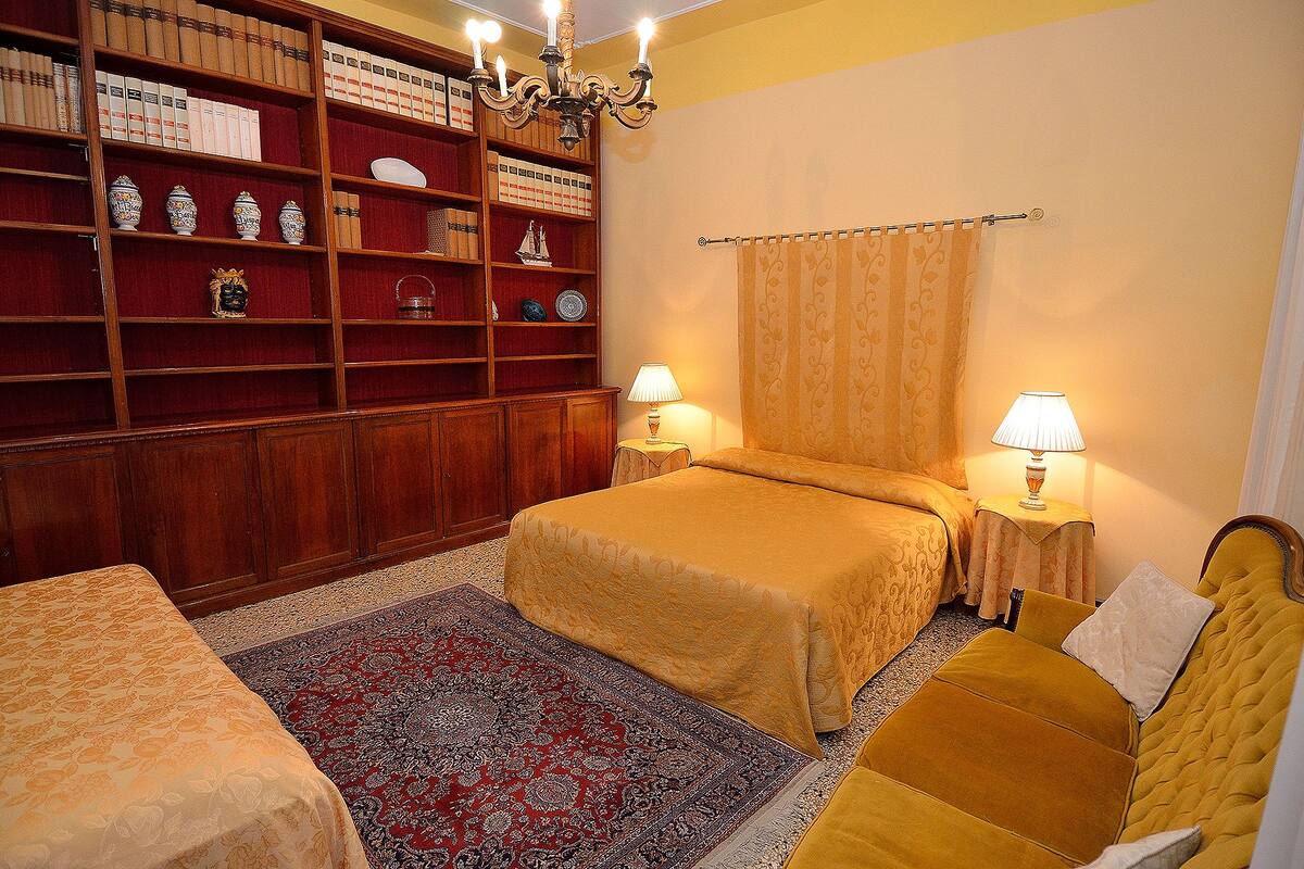 A hotel room with a large wooden bookshelf, two beds with matching headboards, a red and black patterned rug, and two bedside lamps.