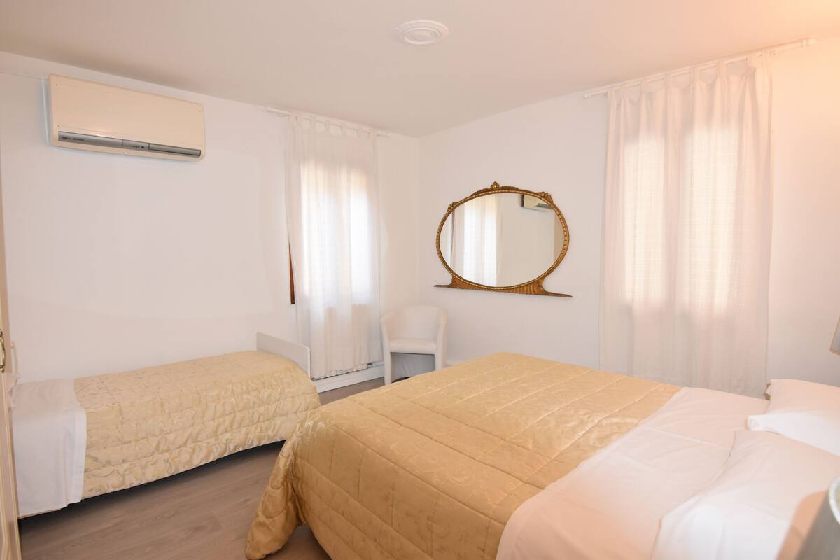 A hotel room with two single beds, a small bedside table, a chair, a round mirror, and an air conditioning unit.