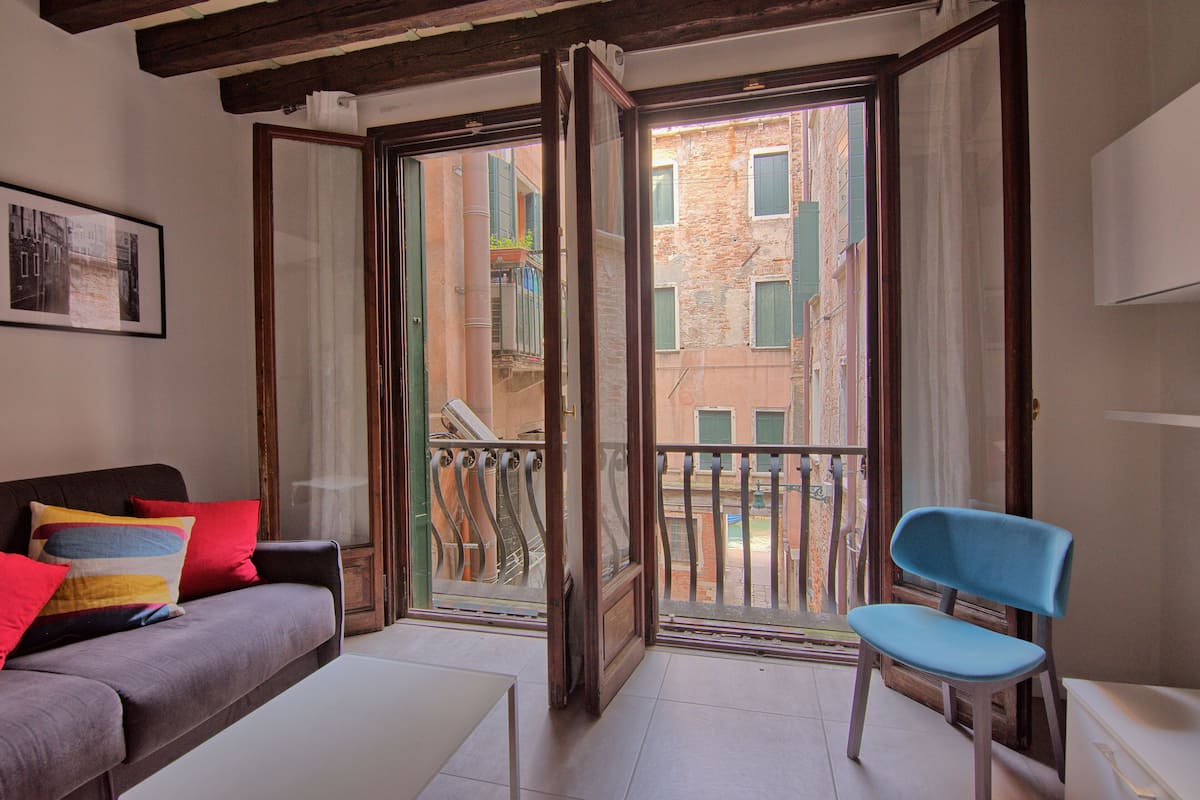 A room with a sofa, a chair, and a balcony with a view of buildings.