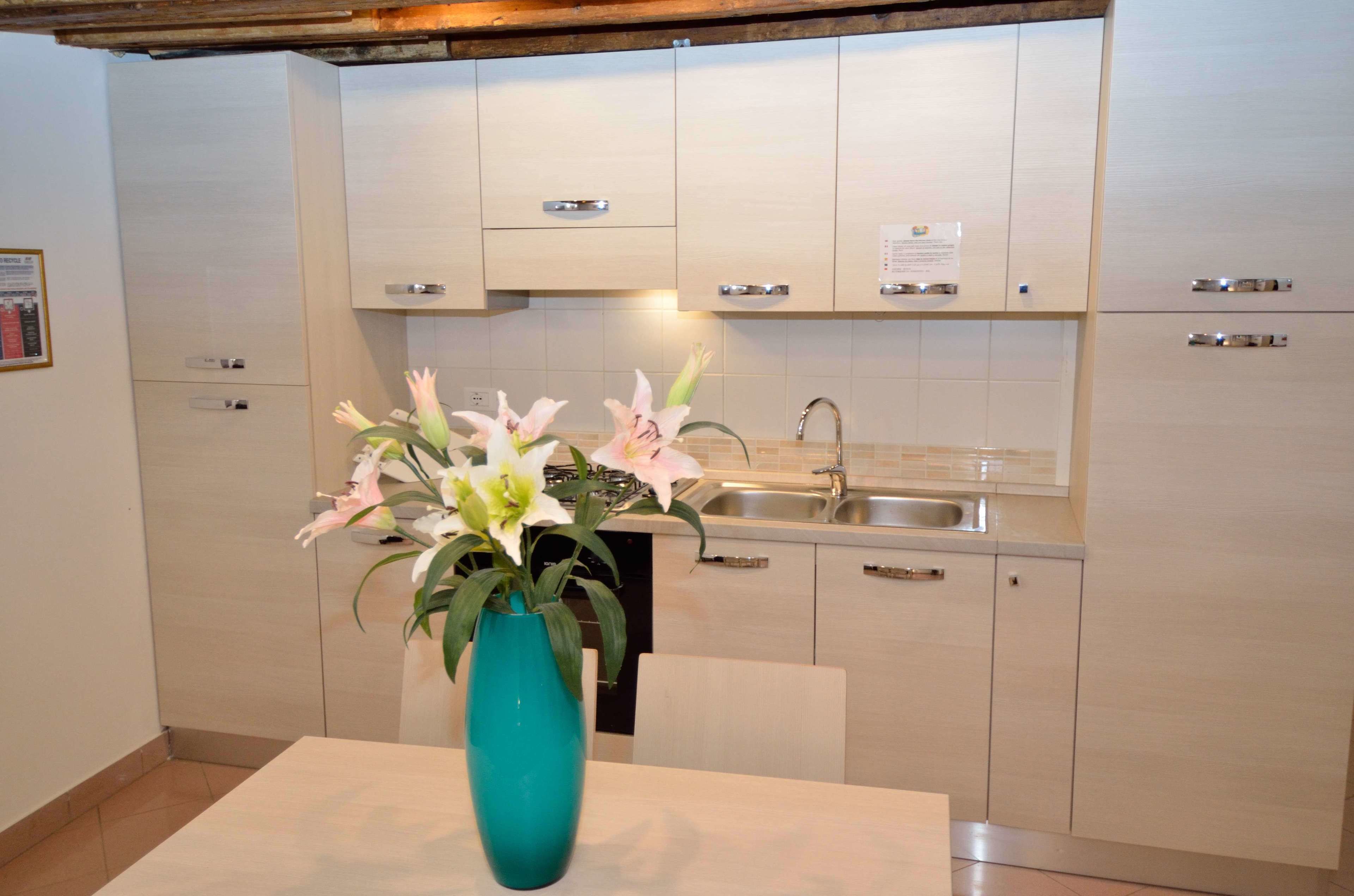 Une cuisine moderne avec des meubles blancs, un évier en acier inoxydable et un vase turquoise orné de fleurs, posé sur le plan de travail.