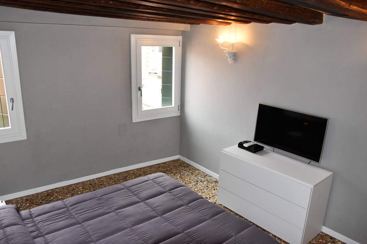 A bedroom with a bed, a TV on a white dresser, a window, and a wall-mounted light fixture.