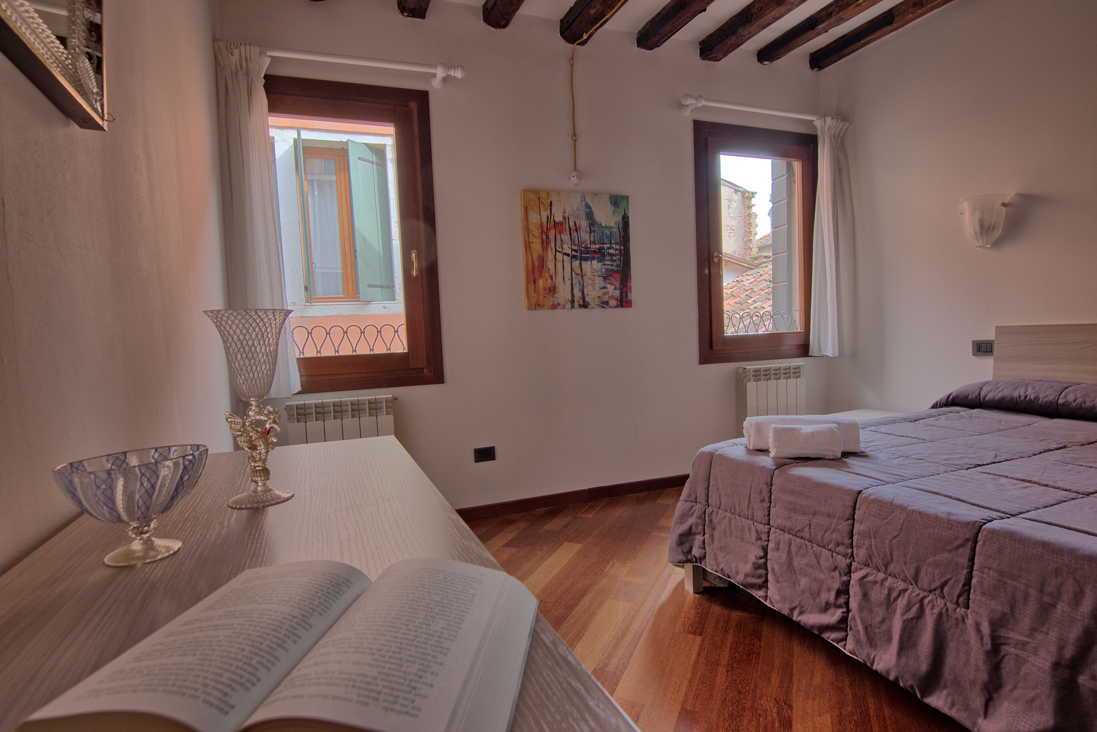 Un dormitorio con una cama, una mesita de noche con un libro y un vaso, dos ventanas con cortinas y un cuadro en la pared.