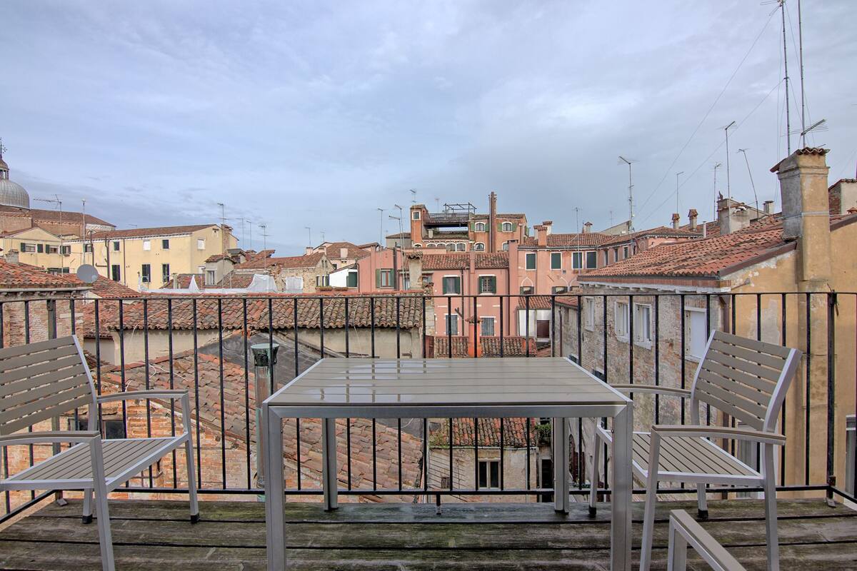 A balcony with a table and two chairs, overlooking a cityscape with terracotta rooftops and historic buildings.