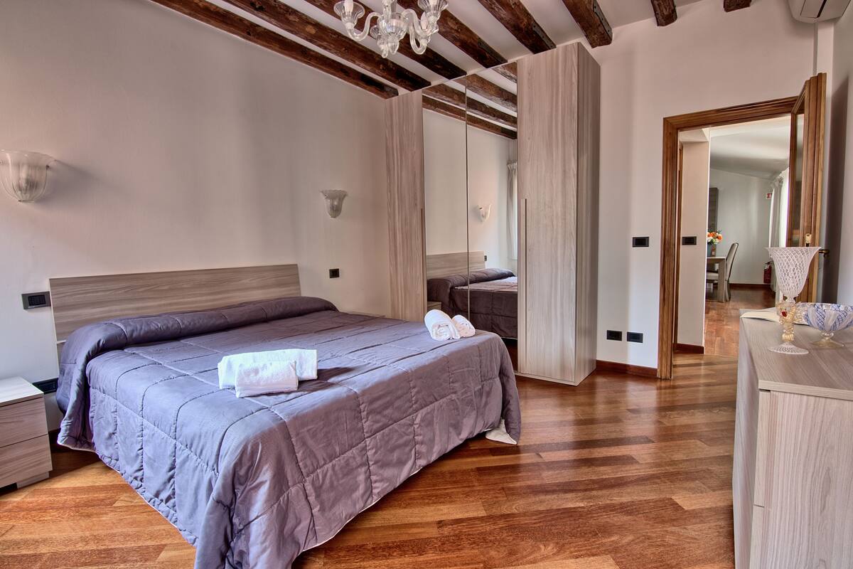 A bedroom with a large bed, wooden beams, a chandelier, a mirror, and a bedside table.