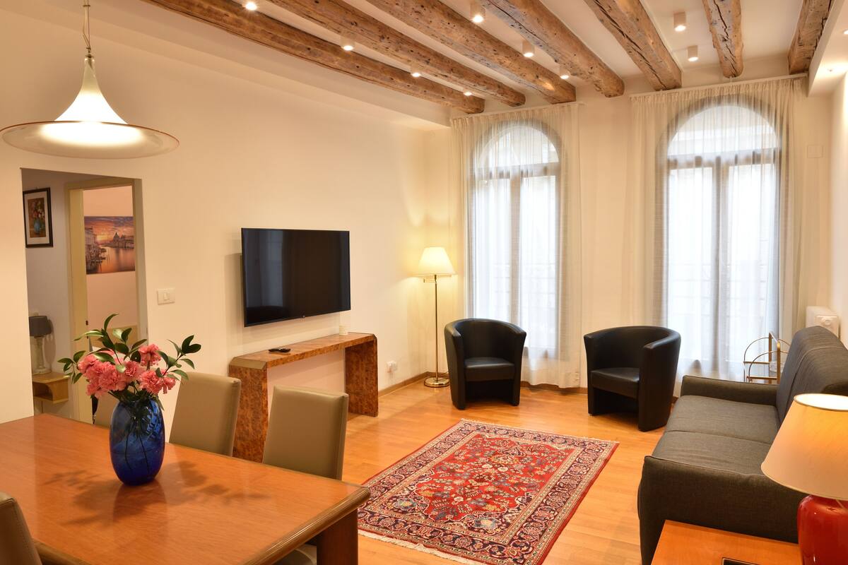 A living room with a TV, sofas, a dining table with chairs, a red rug, and a vase of flowers.