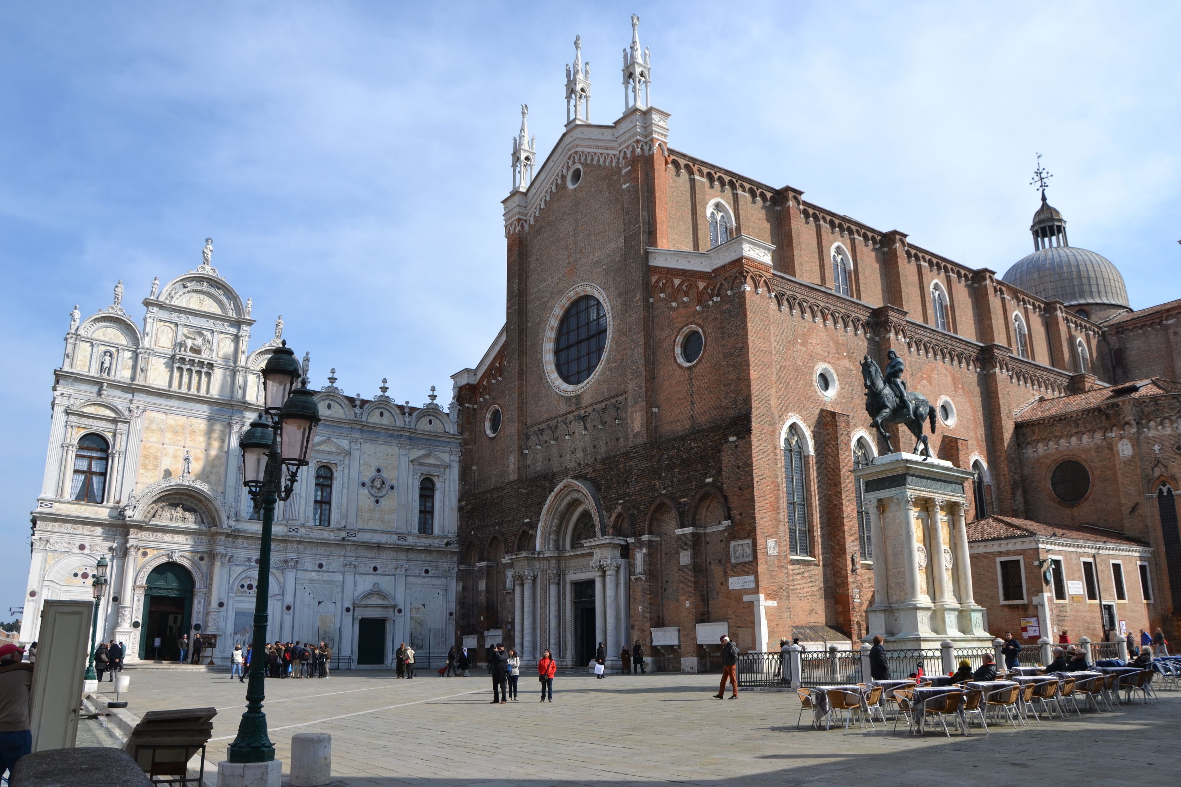 Una plaza histórica con una iglesia grande que tiene una cúpula y estatuas, rodeada de turistas y mesas al aire libre.