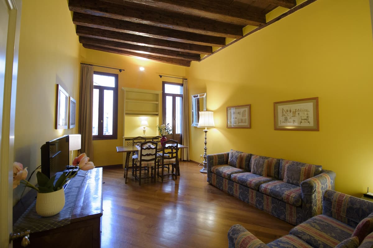A living room with a wooden ceiling, yellow walls, a sofa, a dining table with chairs, and a TV.