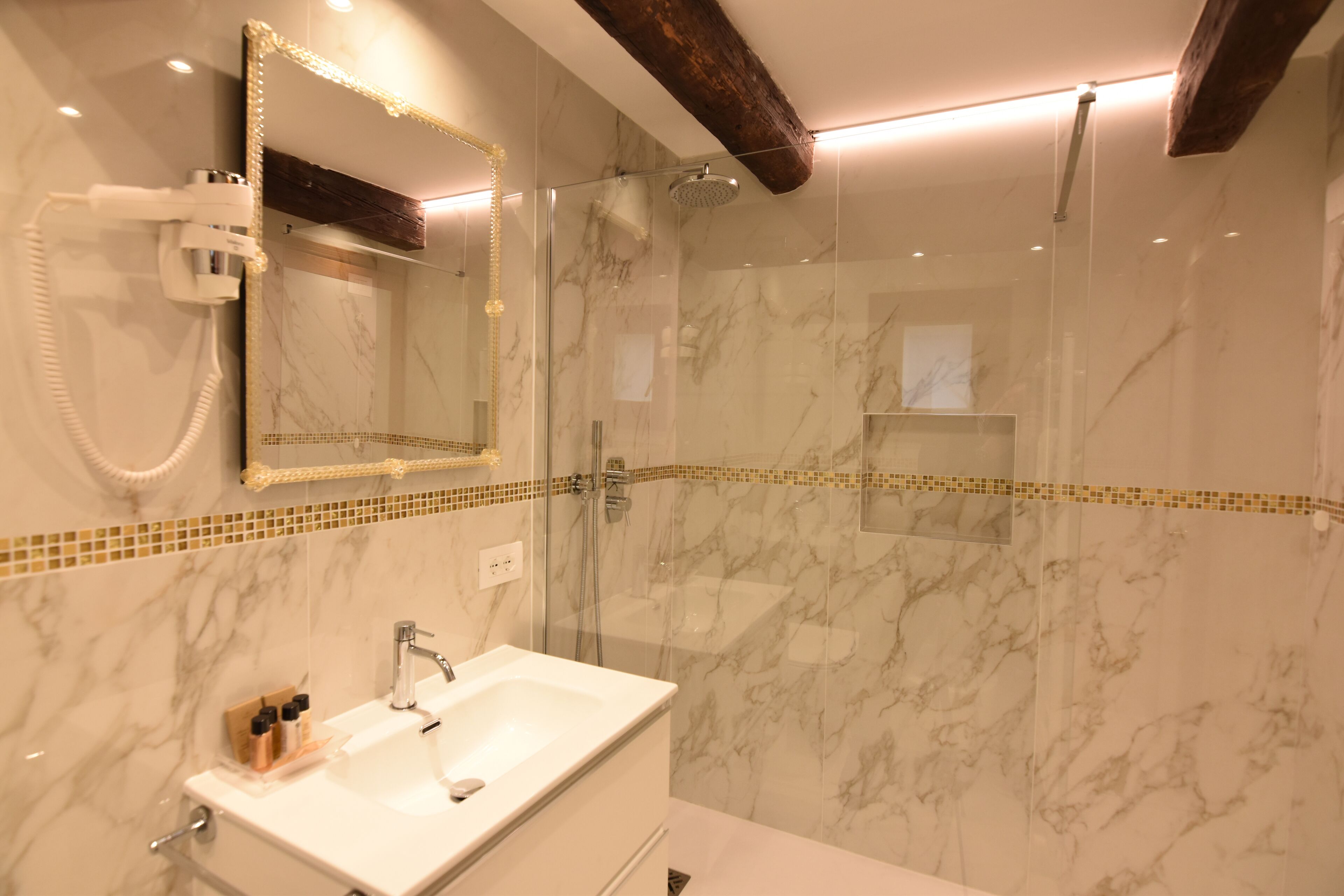 A modern bathroom with a glass-enclosed shower, a marble-tiled wall, and a vanity with a mirror and hairdryer.