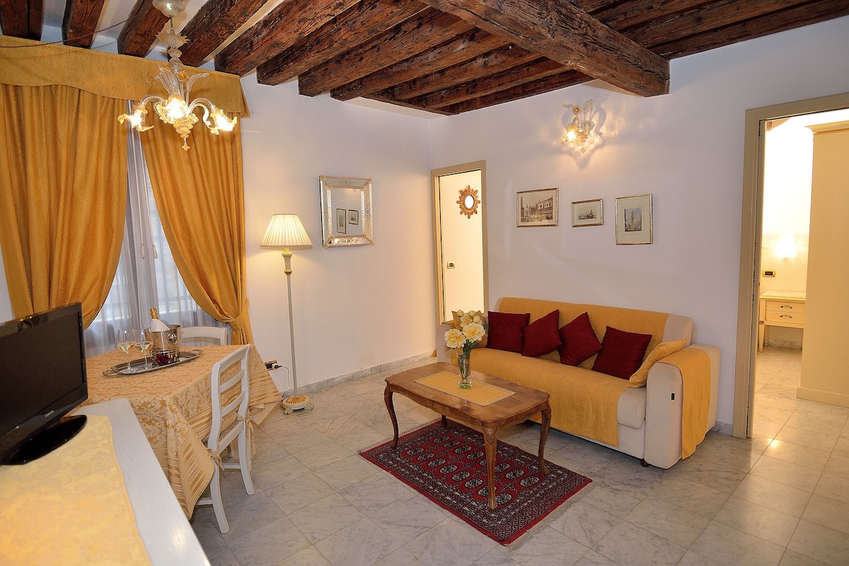 A cozy living room with a sofa, a coffee table, and a TV. There is a dining table with a lamp and a chair. The room has wooden beams on the ceiling and a door with a mirror and pictures on the wall.