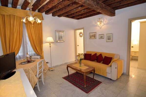 Un salon chaleureux avec un canapé, une table basse et un téléviseur. On y trouve une table à manger avec une lampe et une chaise. La pièce est dotée de poutres en bois au plafond et d’une porte ornée d’un miroir et de tableaux accrochés au mur.