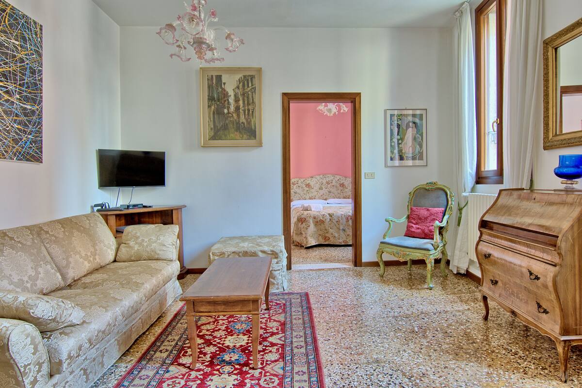 A living room with a sofa, a television, a wooden cabinet, and a bed in the background.