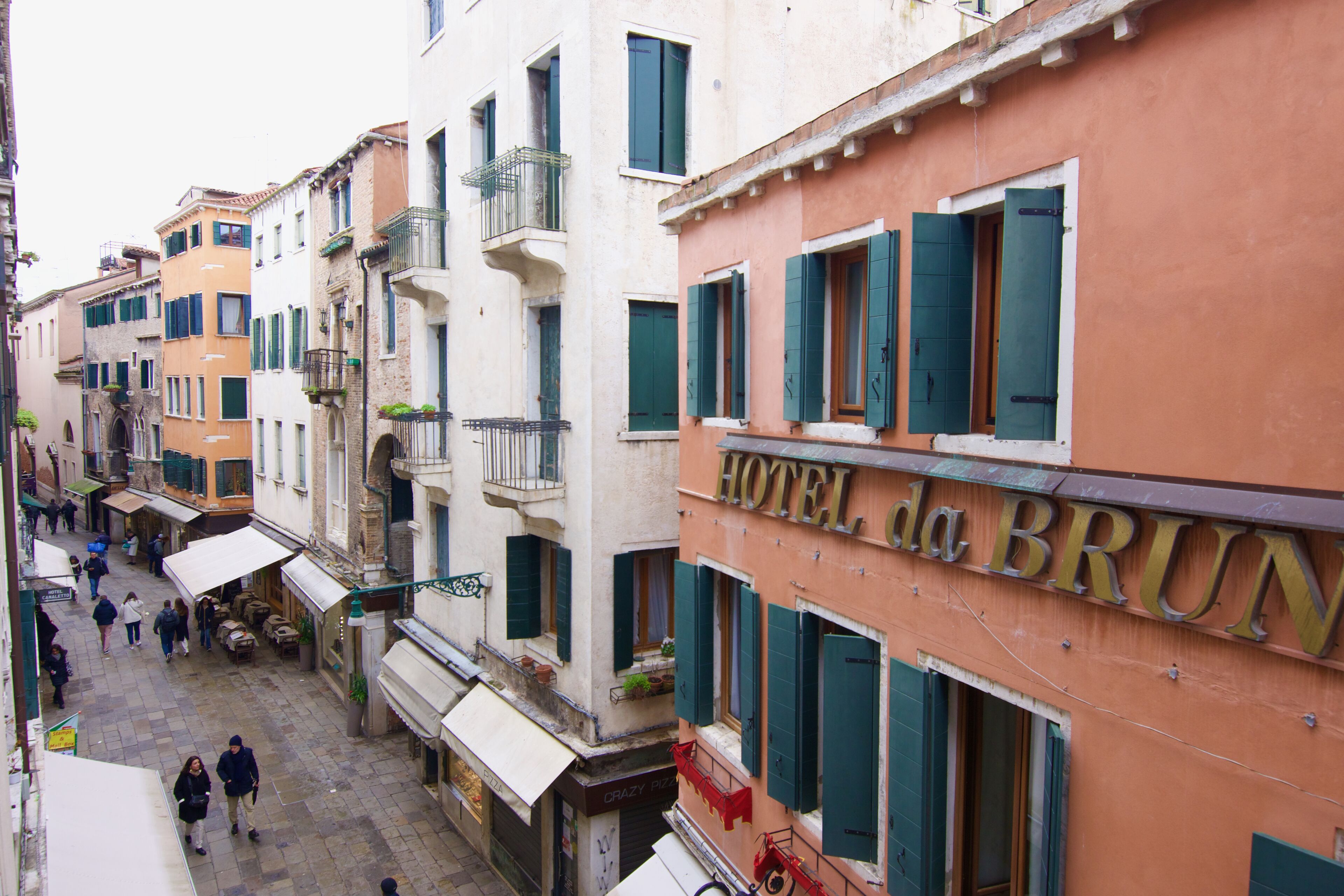 Une vue de la rue où se trouve l’hôtel da Brun, avec une rangée de bâtiments aux volets et balcons verts, une rue pavée et des piétons.