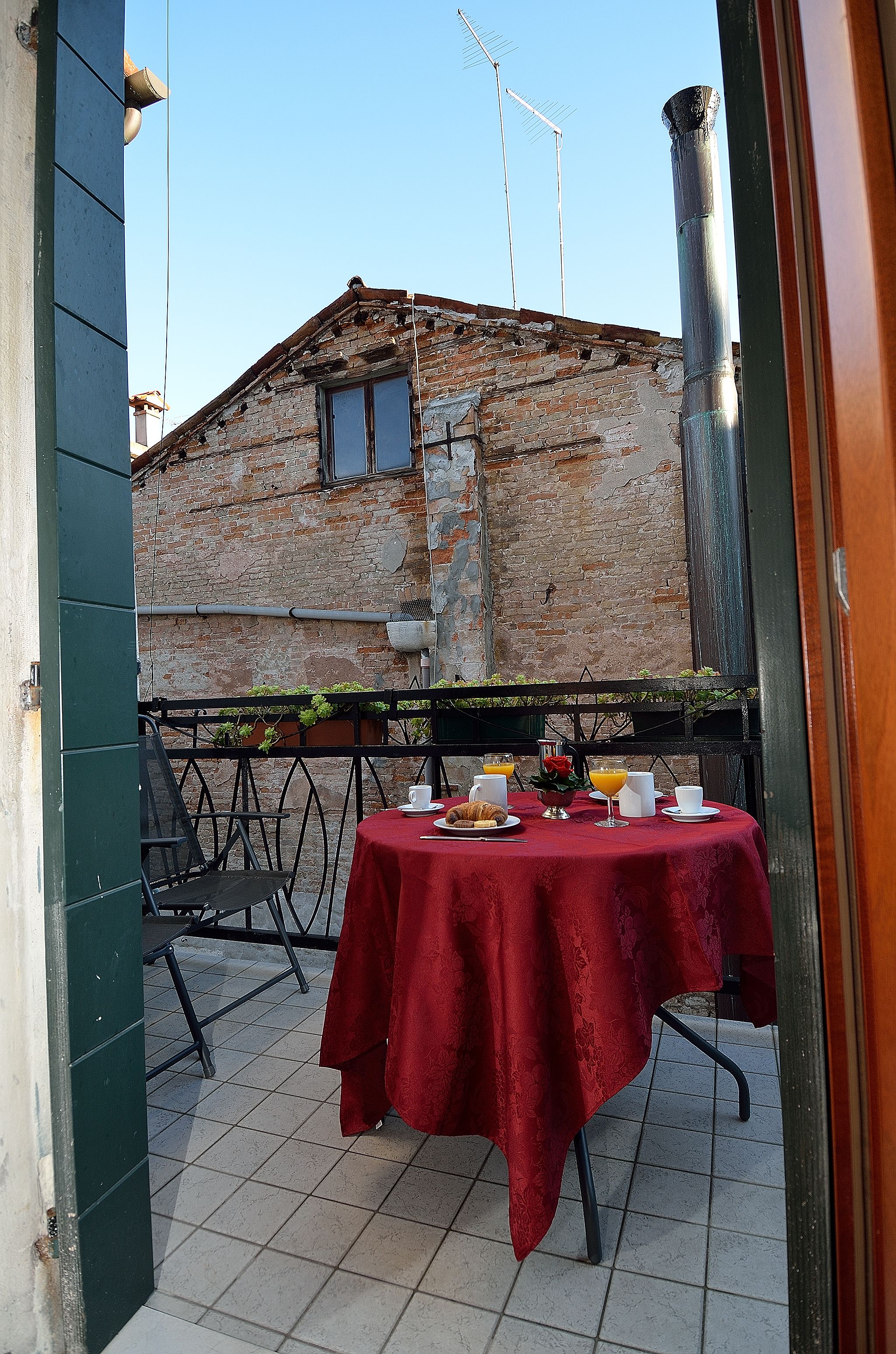 Een terras met een tafel gedekt voor twee, met op de achtergrond een bakstenen gebouw en een metalen hek.