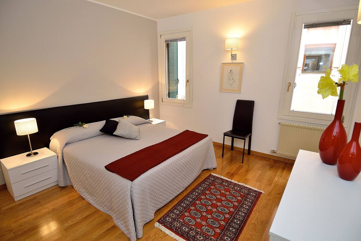A neatly arranged bedroom with a bed, bedside tables, a chair, a window, and a radiator.