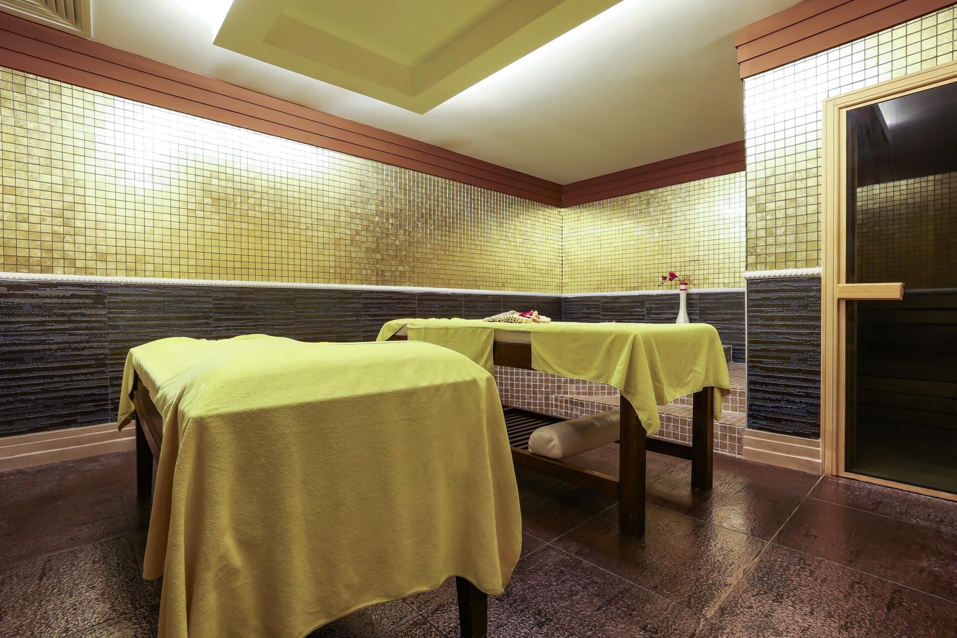 A spa room with two massage tables covered in yellow towels, a tiled wall, and a wooden bench.