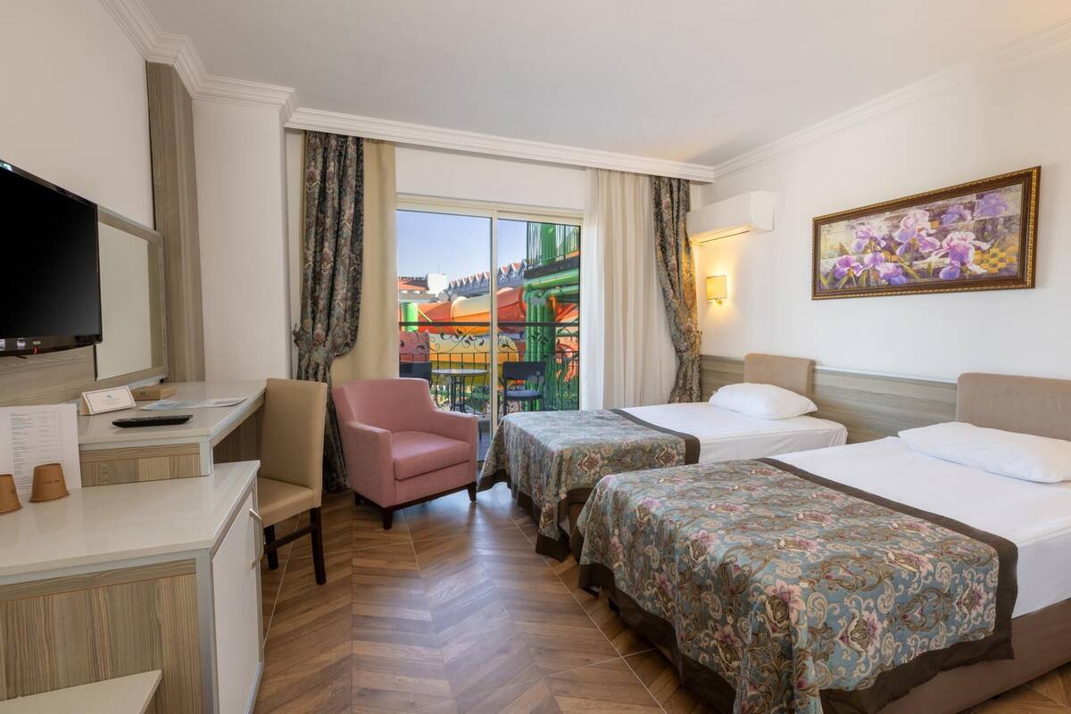 A hotel room with two beds, a desk, a chair, a TV, and a window with a view of colorful buildings.