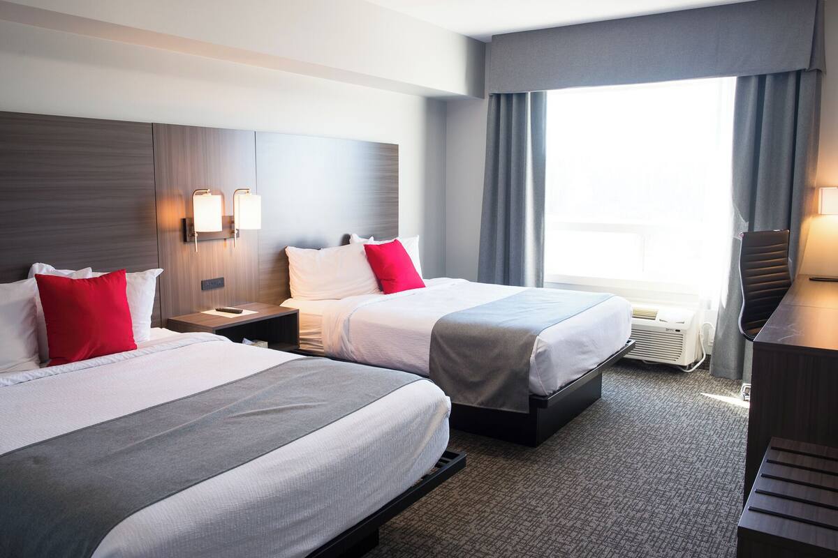A hotel room with two beds, a desk, a chair, a window with curtains, and a wall-mounted air conditioner.