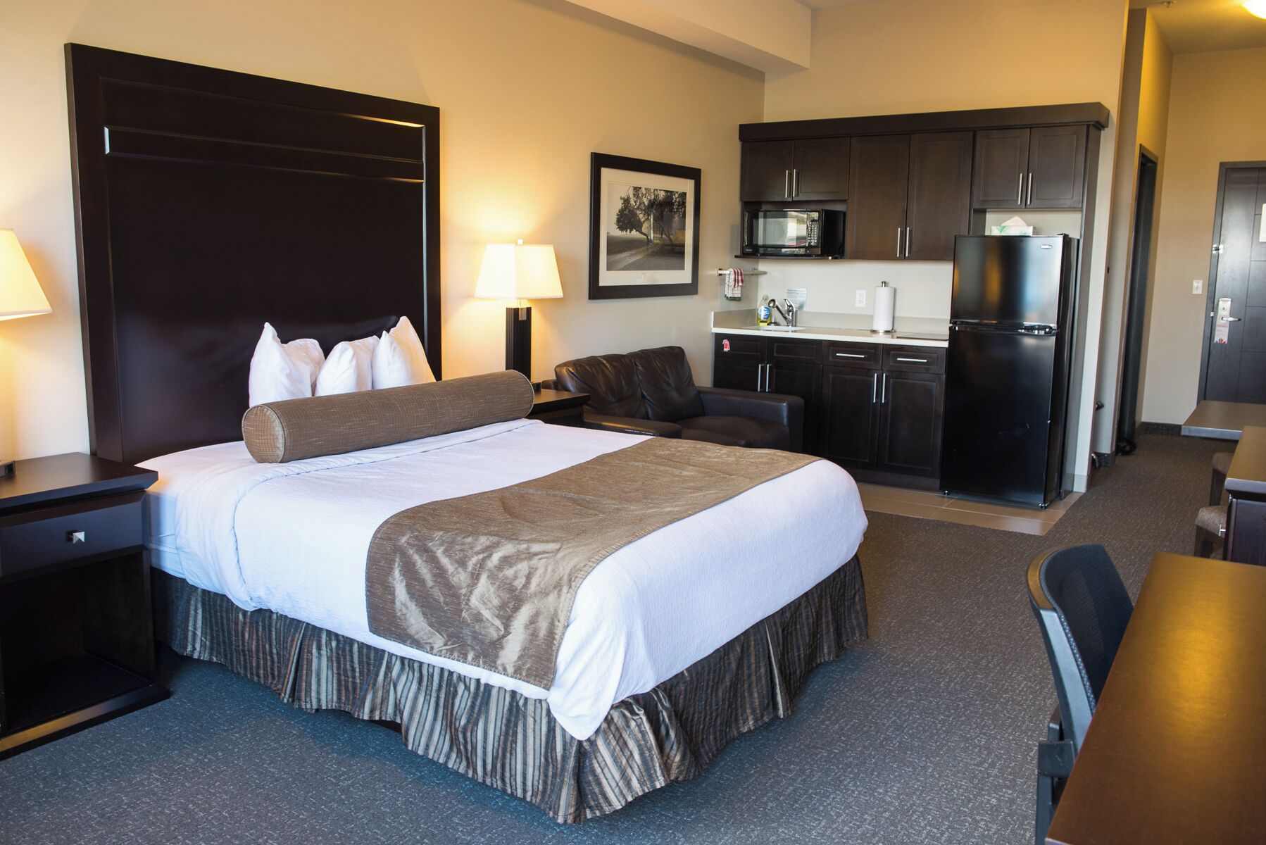Quarto de hotel com uma cama grande, uma mesa, uma cadeira, um sofá, uma kitchenette equipada com frigorífico e micro-ondas, e um quadro na parede.