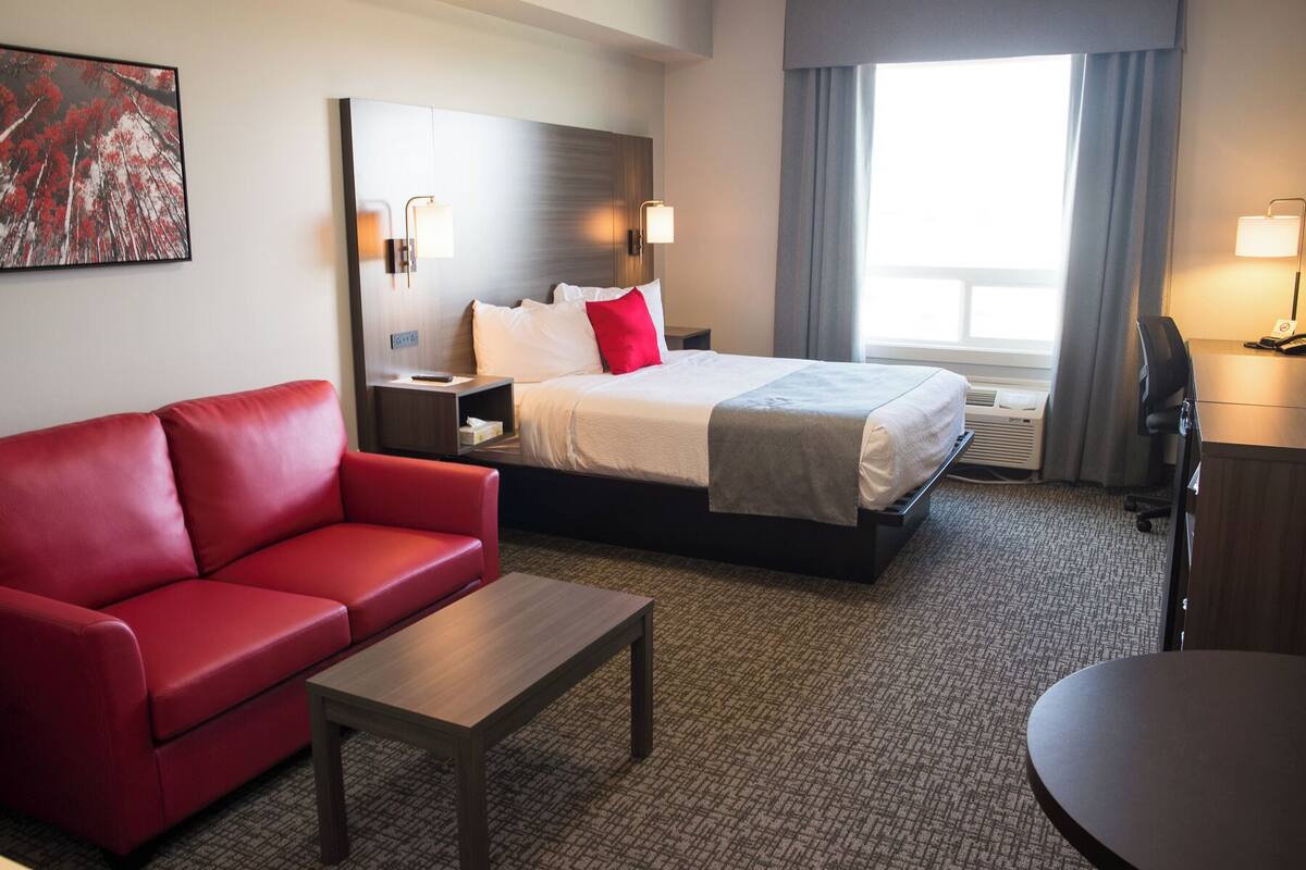 A hotel room with a red sofa, a bed with white and red bedding, a wooden nightstand, a desk, and a large window with curtains.