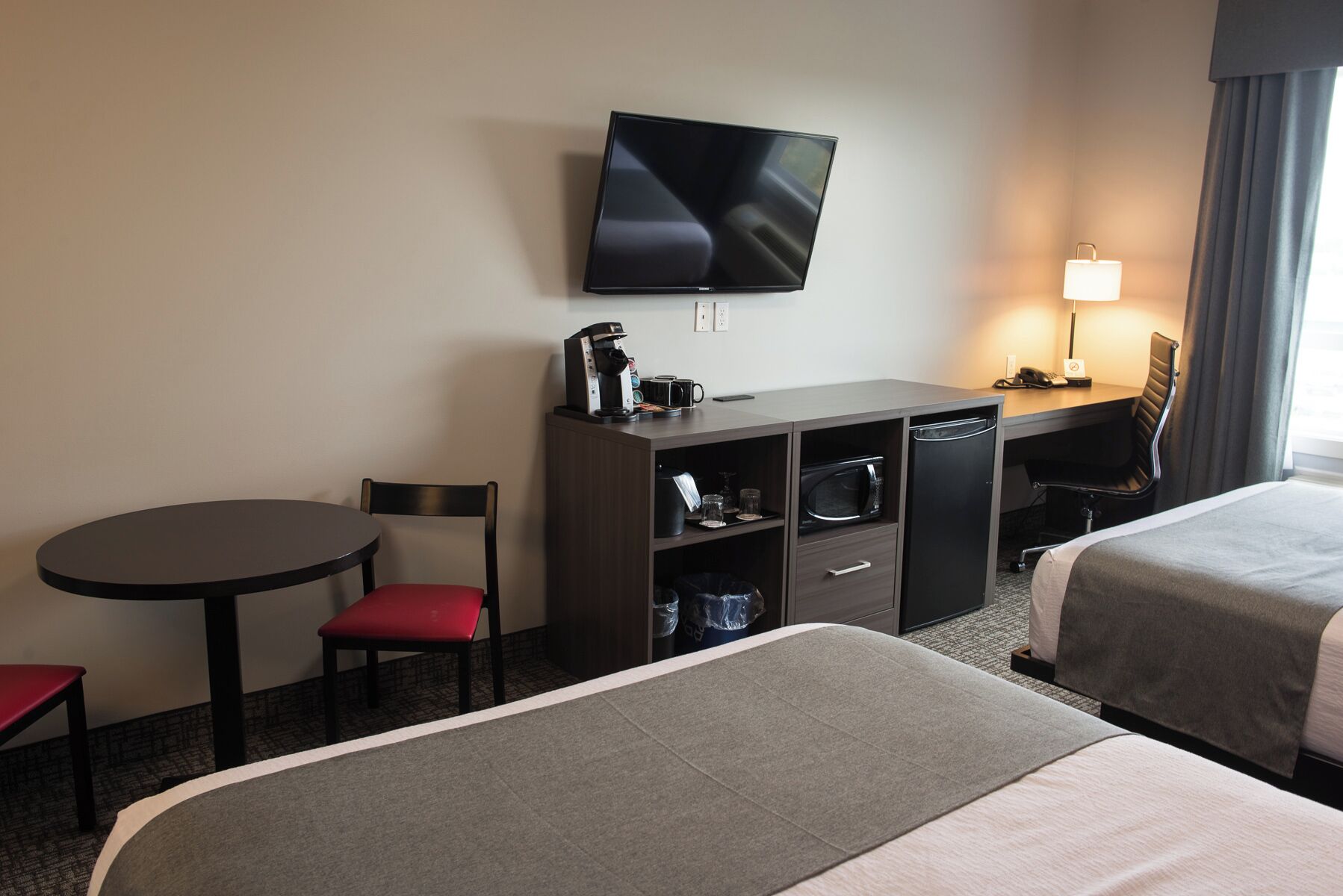 Quarto de hotel com cama, escrivaninha com televisão, cadeira, mesa pequena e abajur.