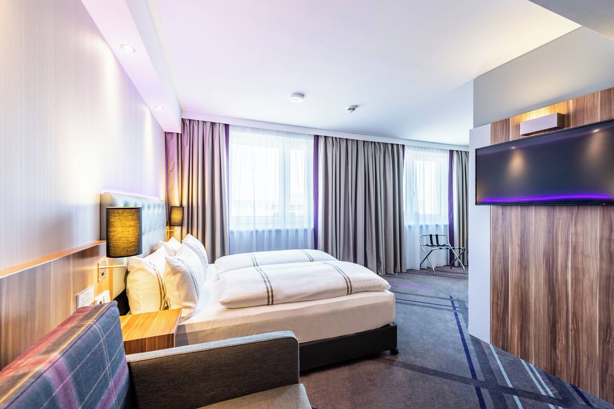 A modern hotel room with a large bed, a desk with a lamp, a flat-screen TV, and a view of the city through large windows.