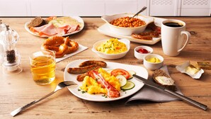A breakfast buffet with various dishes, including eggs, sausages, and toast, set on a wooden table in a kitchen with a blue-tiled backsplash. - Premier Inn Nürnberg City Centre (Nuremberg)