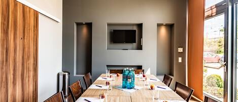 Uma sala de conferências com uma mesa comprida, cadeiras, uma TV de tela plana e uma janela com vista para o exterior.