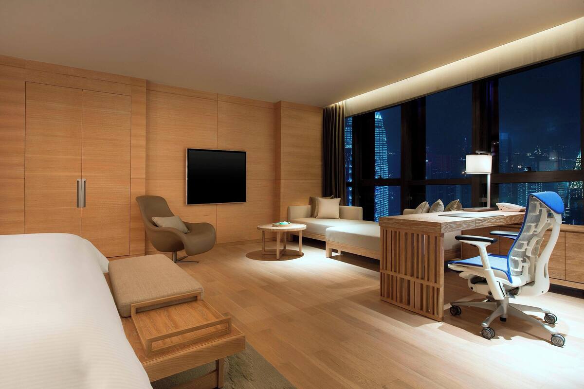 A modern hotel room with a bed, a desk, a chair, a sofa, and a large window with a city view.