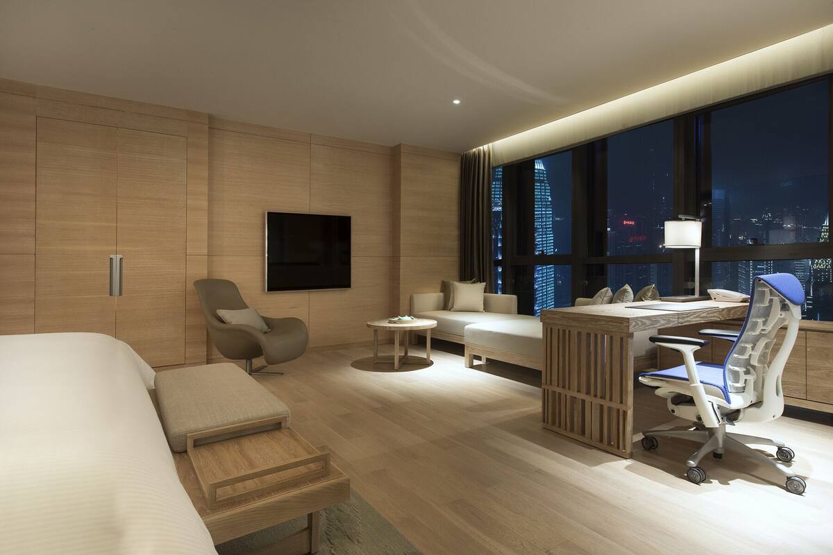 A modern hotel room with a bed, a sofa, a desk with a chair, a TV, and a city view.