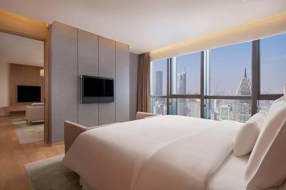 A modern hotel room with a large bed, a flat-screen TV mounted on the wall, and a city view through large windows.