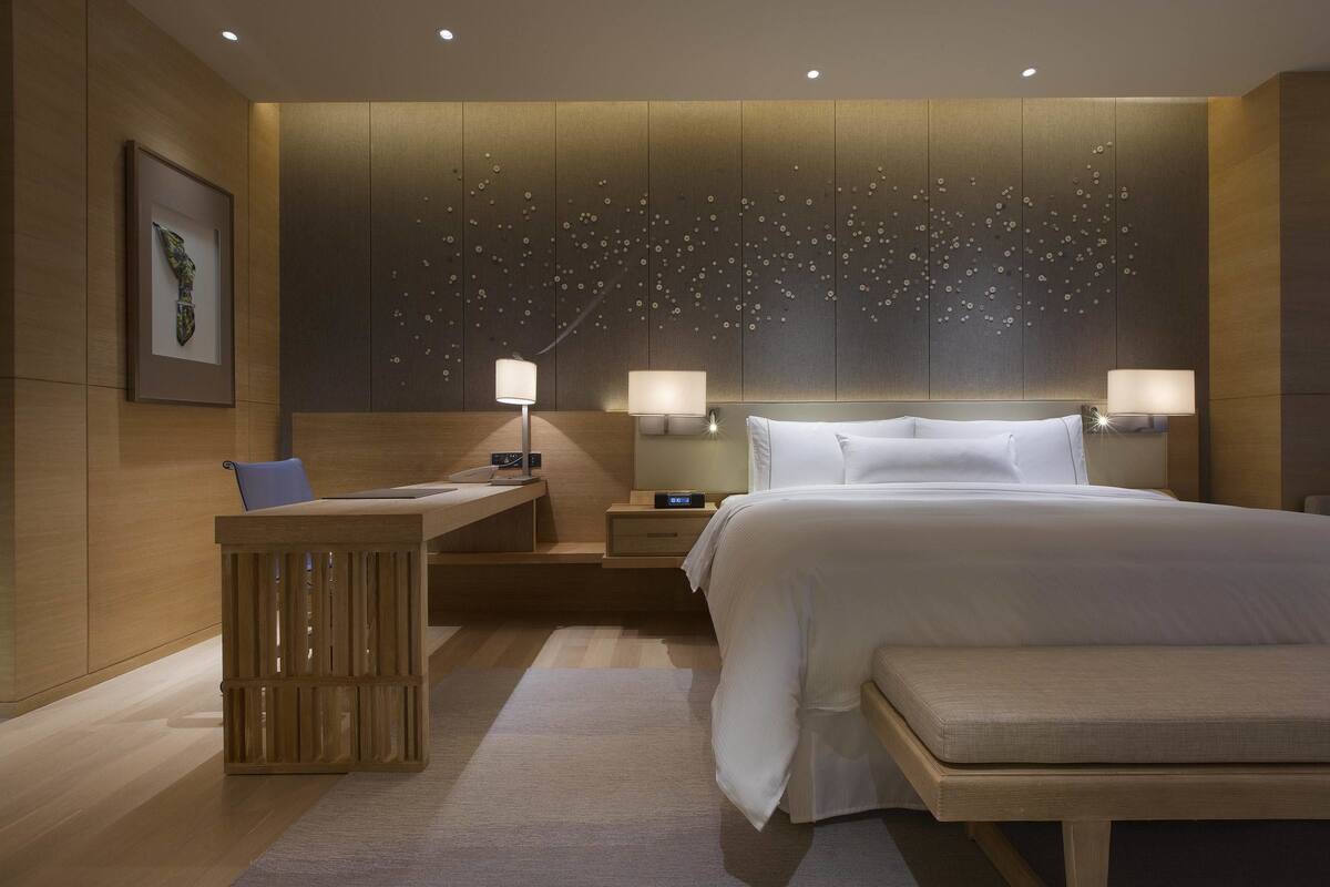 A modern hotel room with a large bed, bedside tables with lamps, a desk with a chair, and a decorative wall panel.