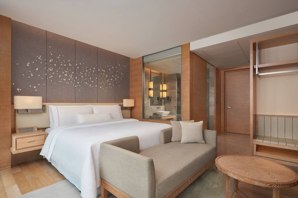 A modern hotel room with a large bed, a sofa, a small table, a bathroom with a mirror and sink, and a wooden wall panel with decorative elements.