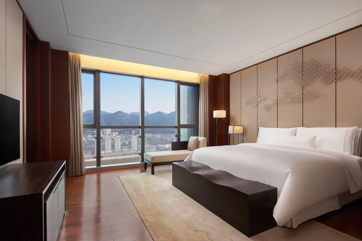 A modern hotel room with a large bed, a flat-screen TV, a seating area, and a view of the city and mountains.