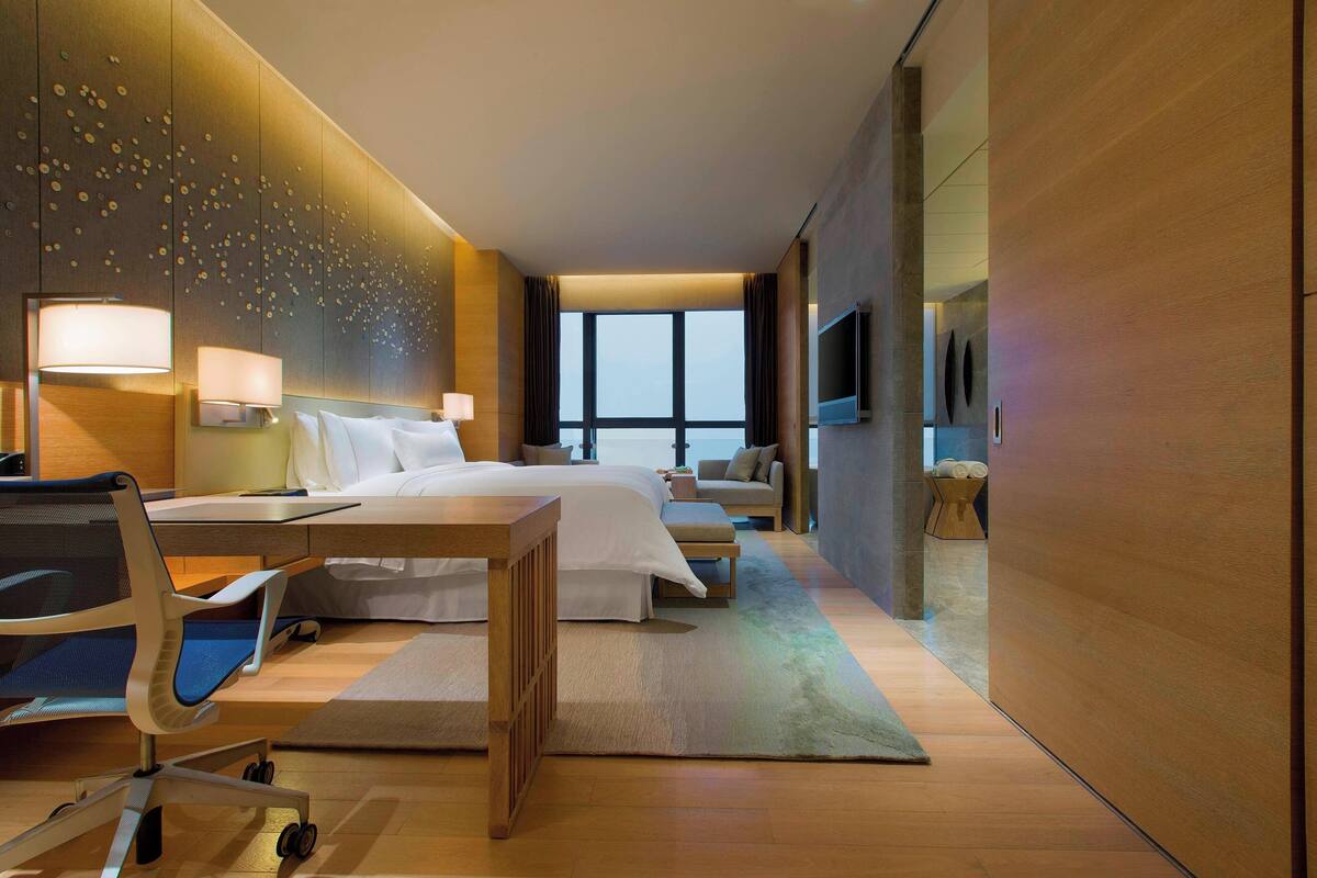 A modern hotel room with a large bed, a desk, a chair, a TV, and a large window.