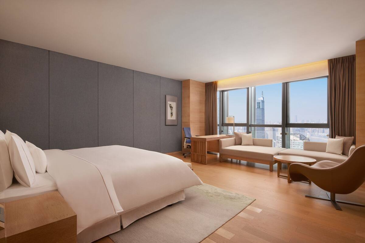 A modern hotel room with a large bed, a desk, a sofa, and a view of the city skyline.