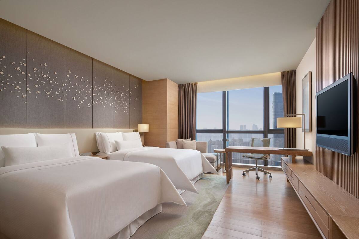 A hotel room with two beds, a desk, a TV, and a city view.