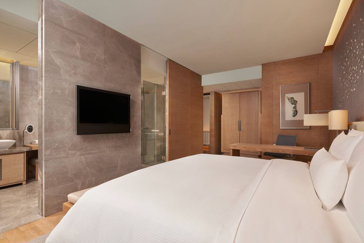 A modern hotel room with a large bed, a flat-screen TV, a desk, and a bathroom with a glass shower.
