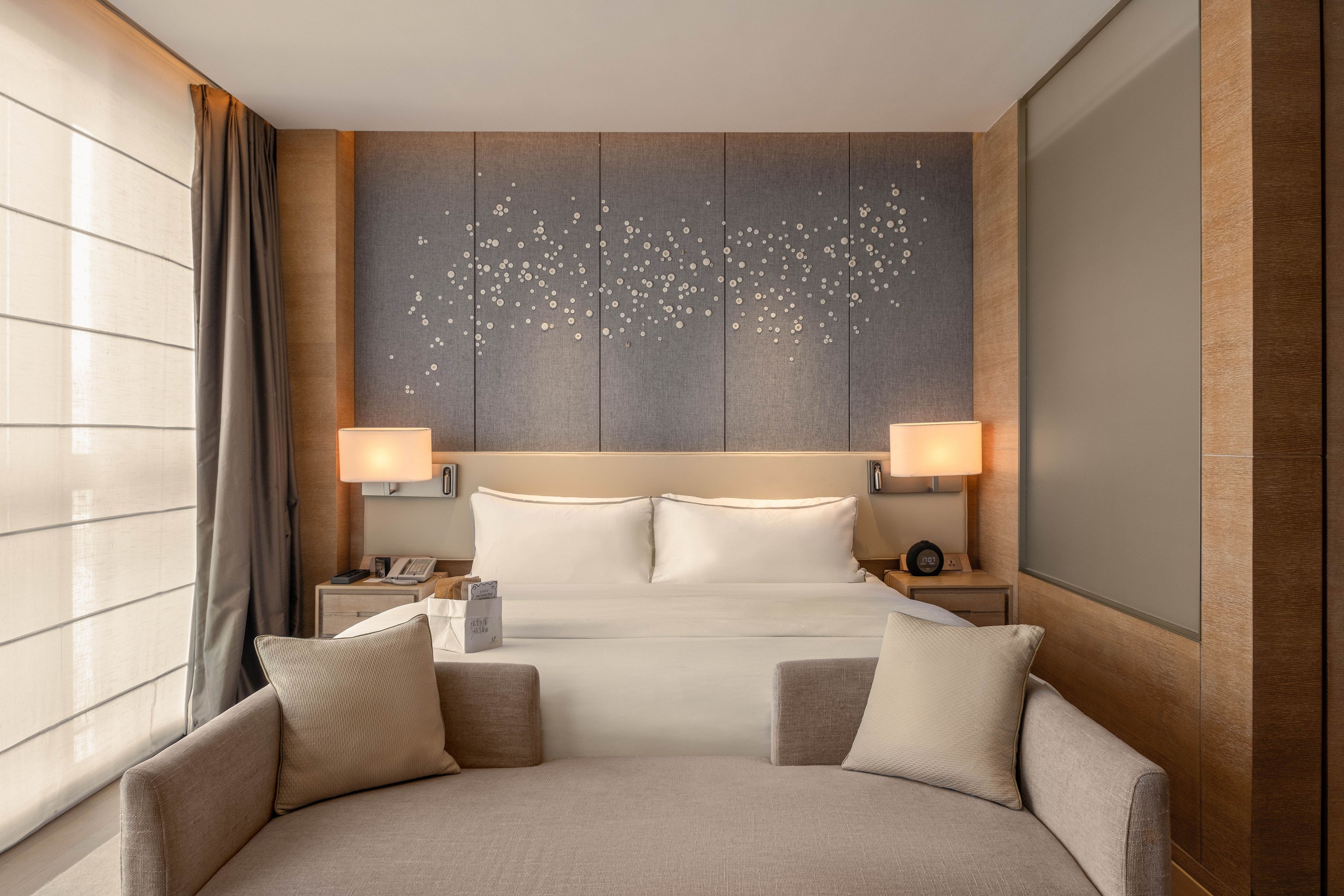 A hotel room with a large bed, two bedside tables, a nightstand, a sofa, and a window with curtains.