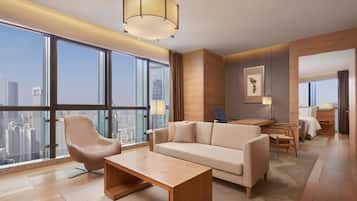 A modern hotel room with a large window offering a city view, a sofa, a coffee table, and a desk with a chair.
