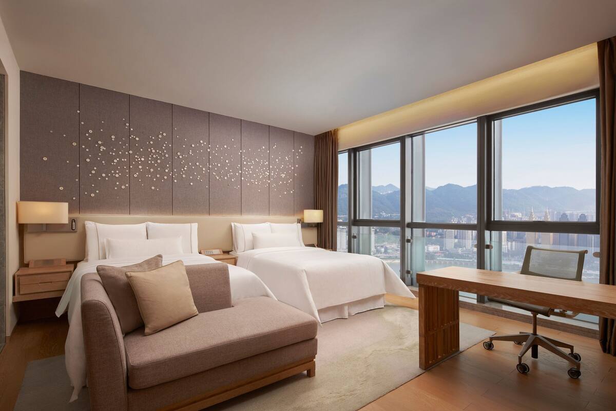 A modern hotel room with two beds, a desk, and a large window offering a city view.