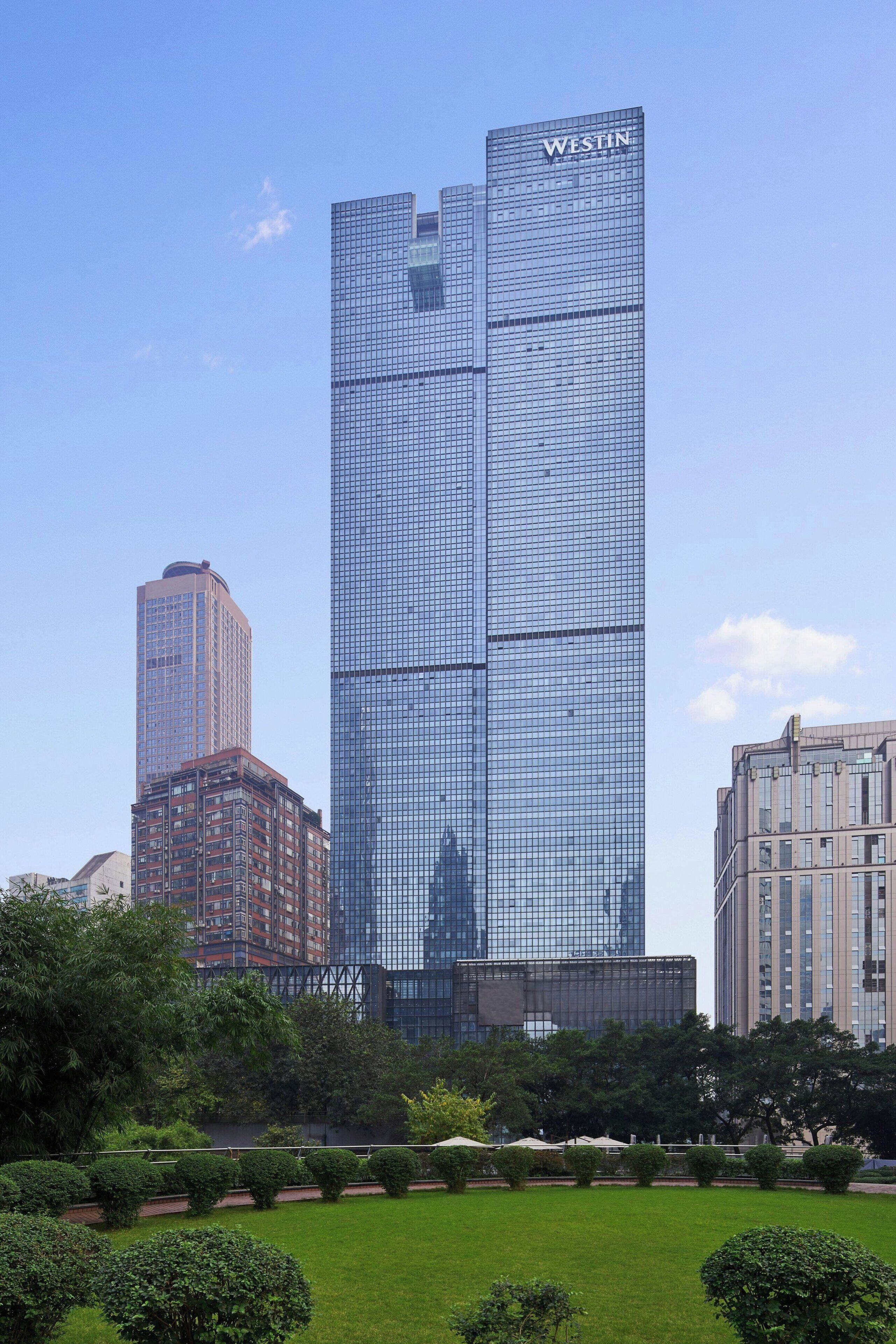 A tall, modern skyscraper with the name "WESTIN" on its facade, surrounded by a well-maintained park with manicured lawns and trees.