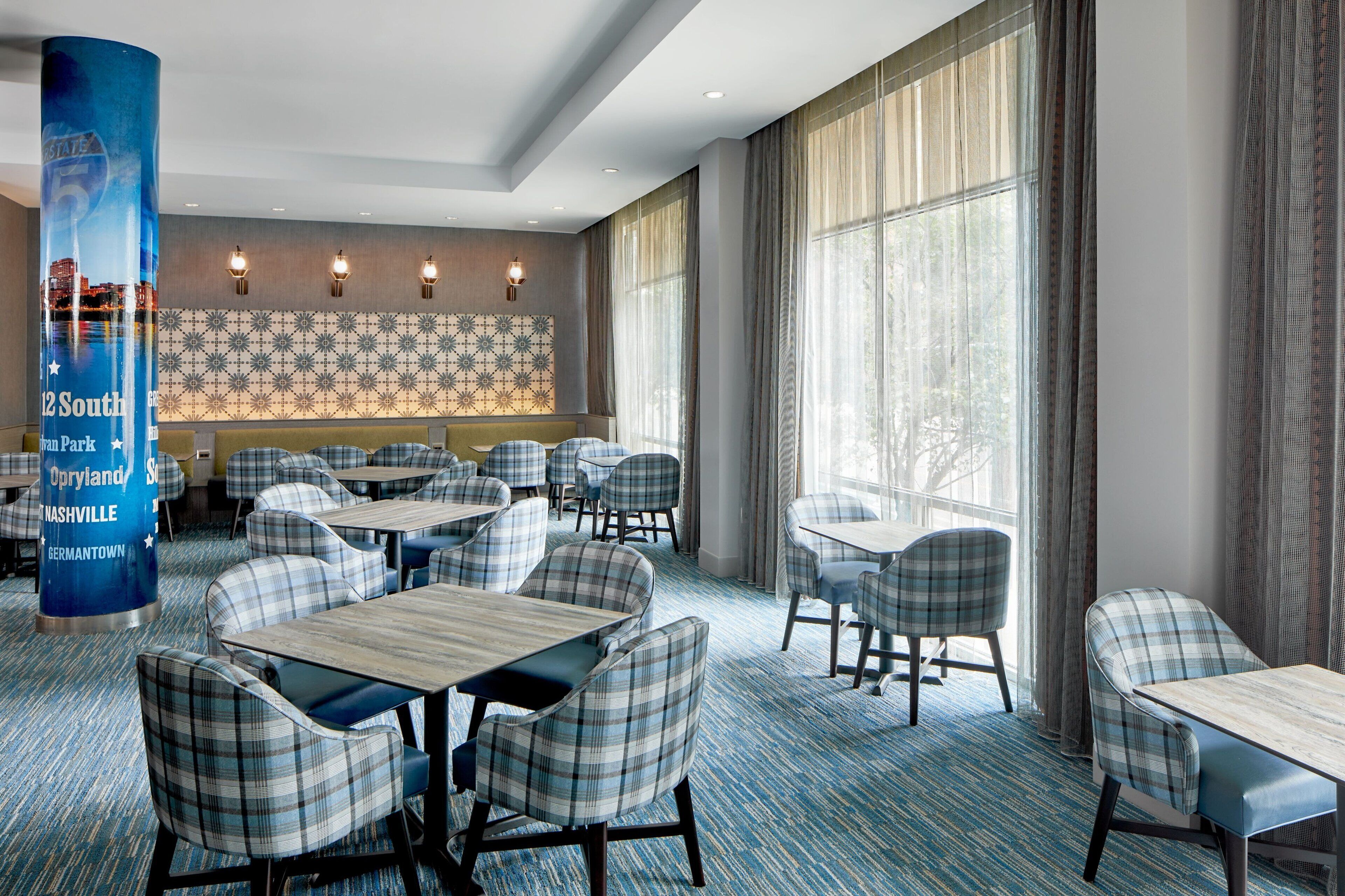 A hotel lobby with a blue carpet, plaid chairs, and tables. There is a large pillar with a cityscape image and text.