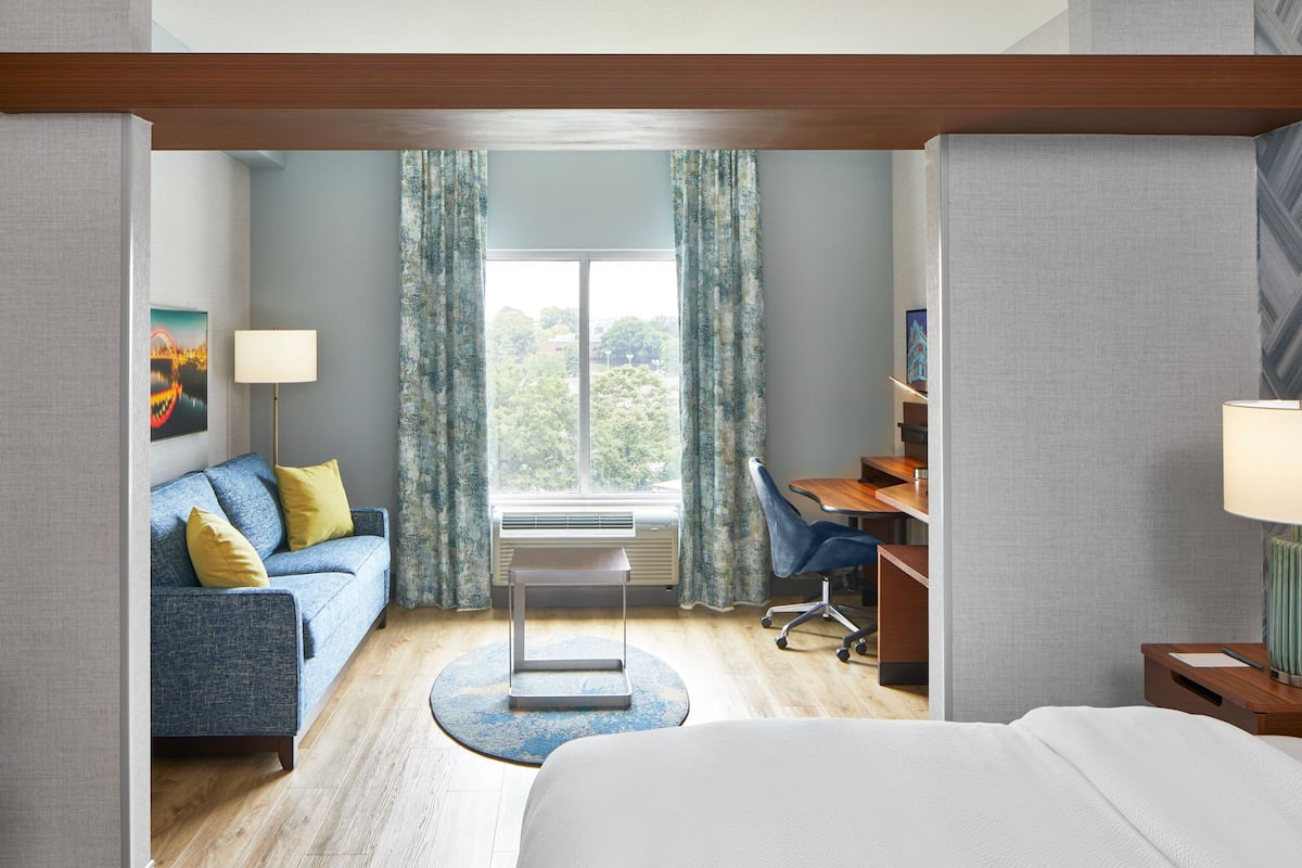 Una habitación de hotel con una cama, un escritorio con una silla, un sofá con cojines, una lámpara de pie y vistas al exterior a través de una ventana.