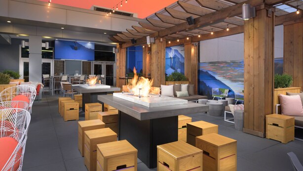 An outdoor seating area with wooden stools, a central fire feature, and a view of a mountainous landscape.