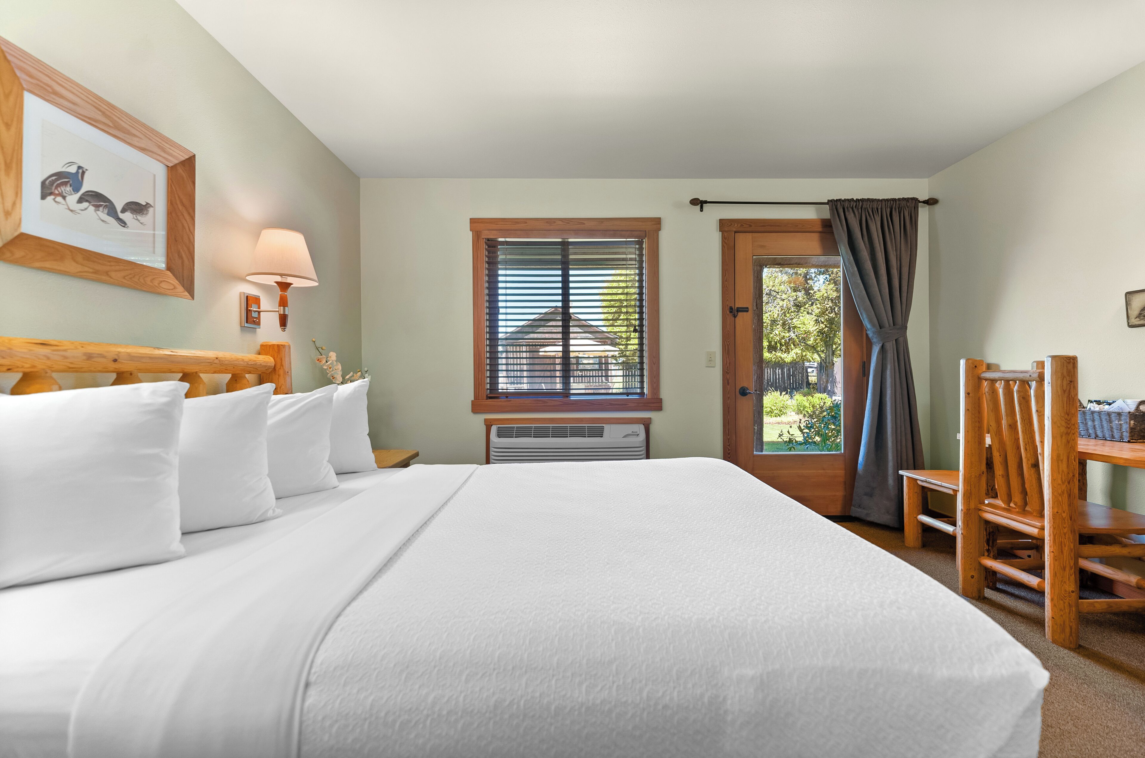 A neatly made bed with white linens, a wooden headboard, a wall-mounted lamp, a window with blinds, a door with a curtain, and a wooden chair.
