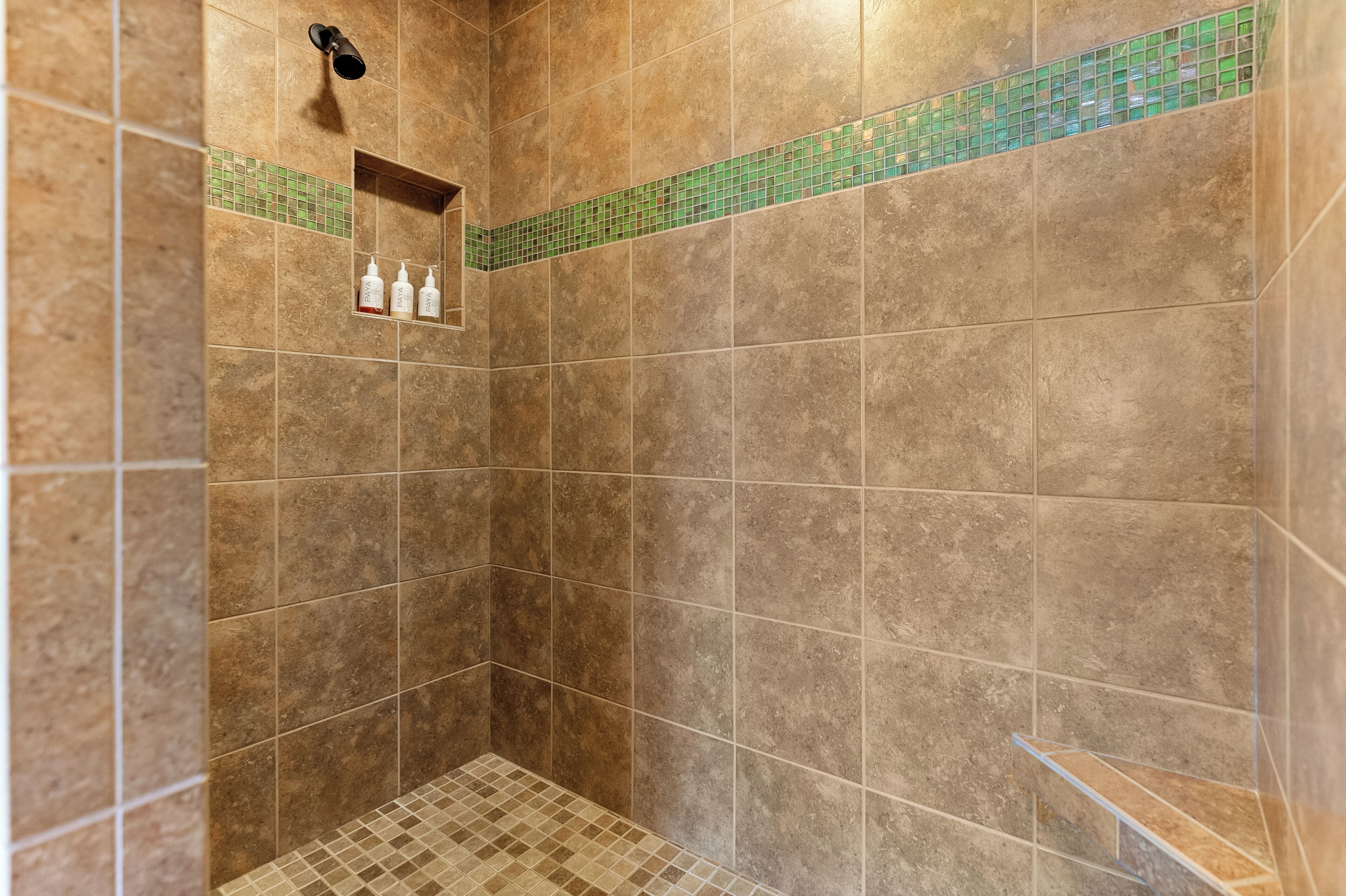 A shower area with beige tiles, a green mosaic border, and a shelf with toiletries.