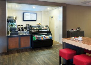 Ein Frühstücksbuffet mit einer gekühlten Verkaufstheke, Kaffeeautomaten und Sitzgelegenheiten mit roten Kissen.