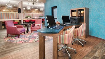 A hotel lobby with a reception desk, two computers, a bookshelf, and a seating area with chairs.