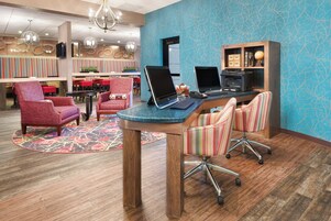 Um saguão de hotel com uma mesa de recepção, dois computadores, uma estante de livros e uma área de estar com cadeiras.