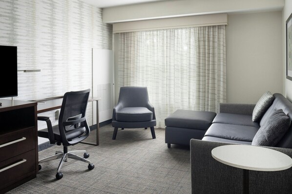 A hotel room with a desk, a black office chair, a grey sofa, and a small round table.
